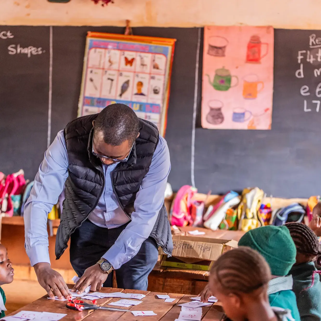Students in light blue uniforms sitting at wooden desks in a classroom with a teacher standing by the window